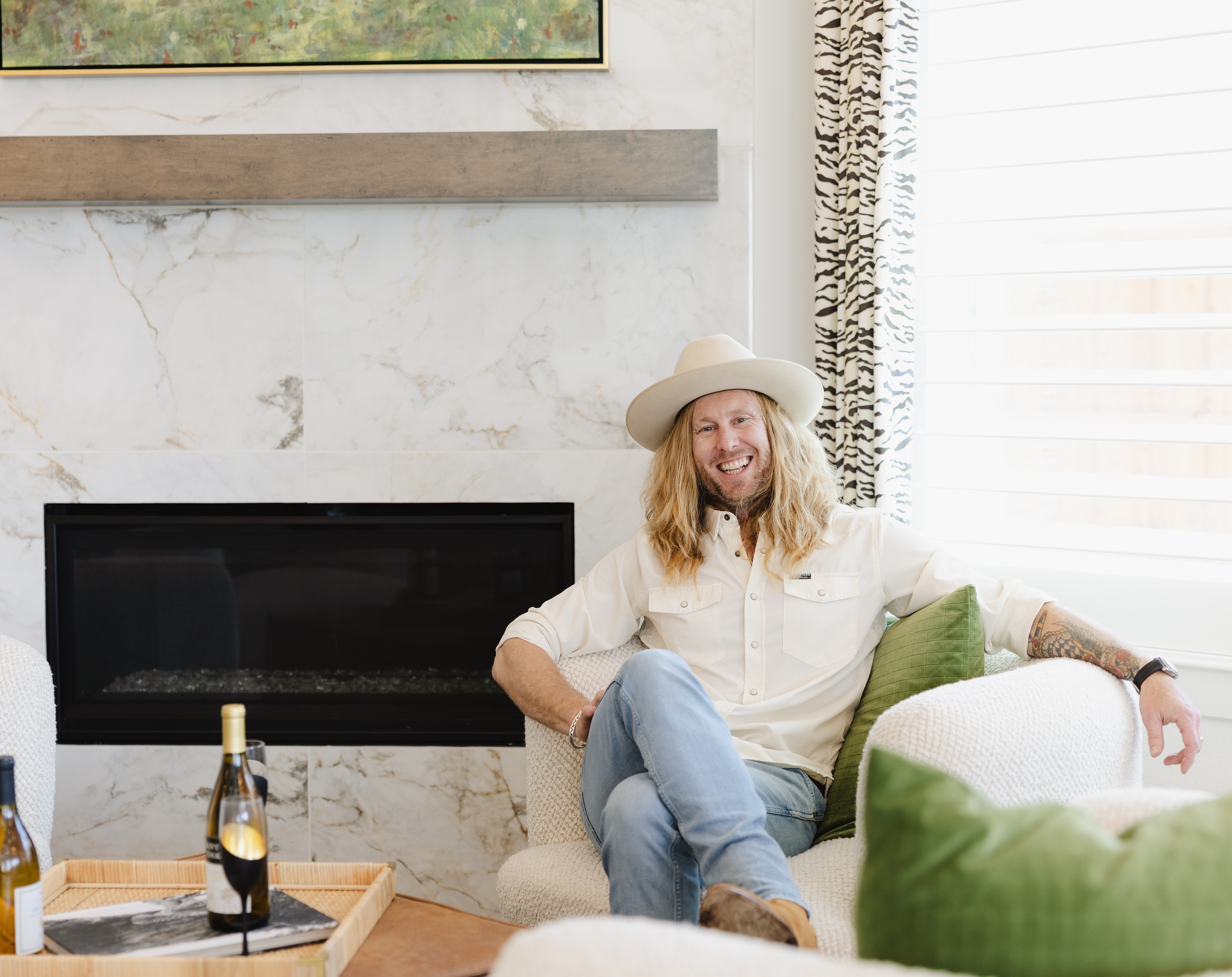 Jamey Ice laughing on a sofa beside a marble fireplace in a finished living room, in a white shirt and hat.