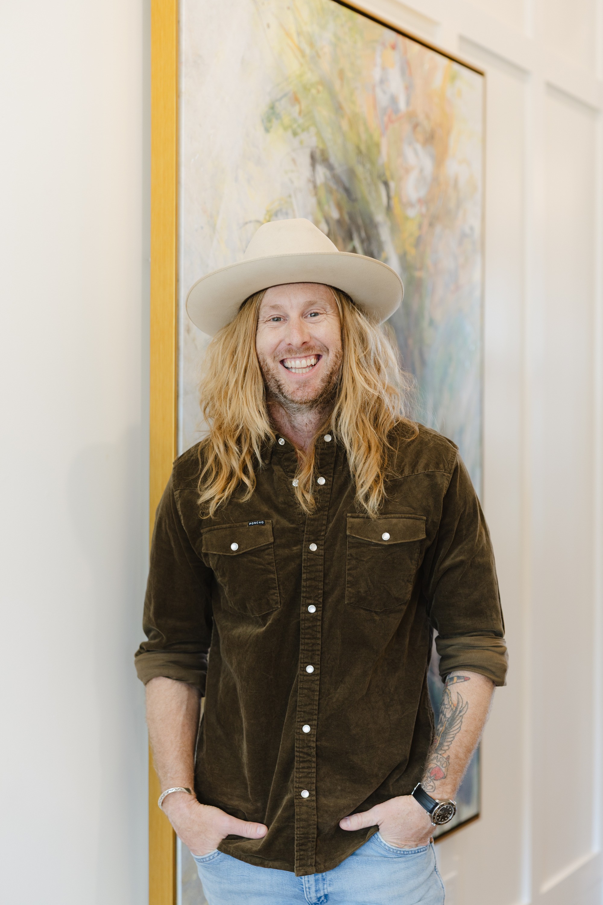 Jamey Ice laughing with his hands in his pockets in a hallway, a tall painting with a gold frame behind him.