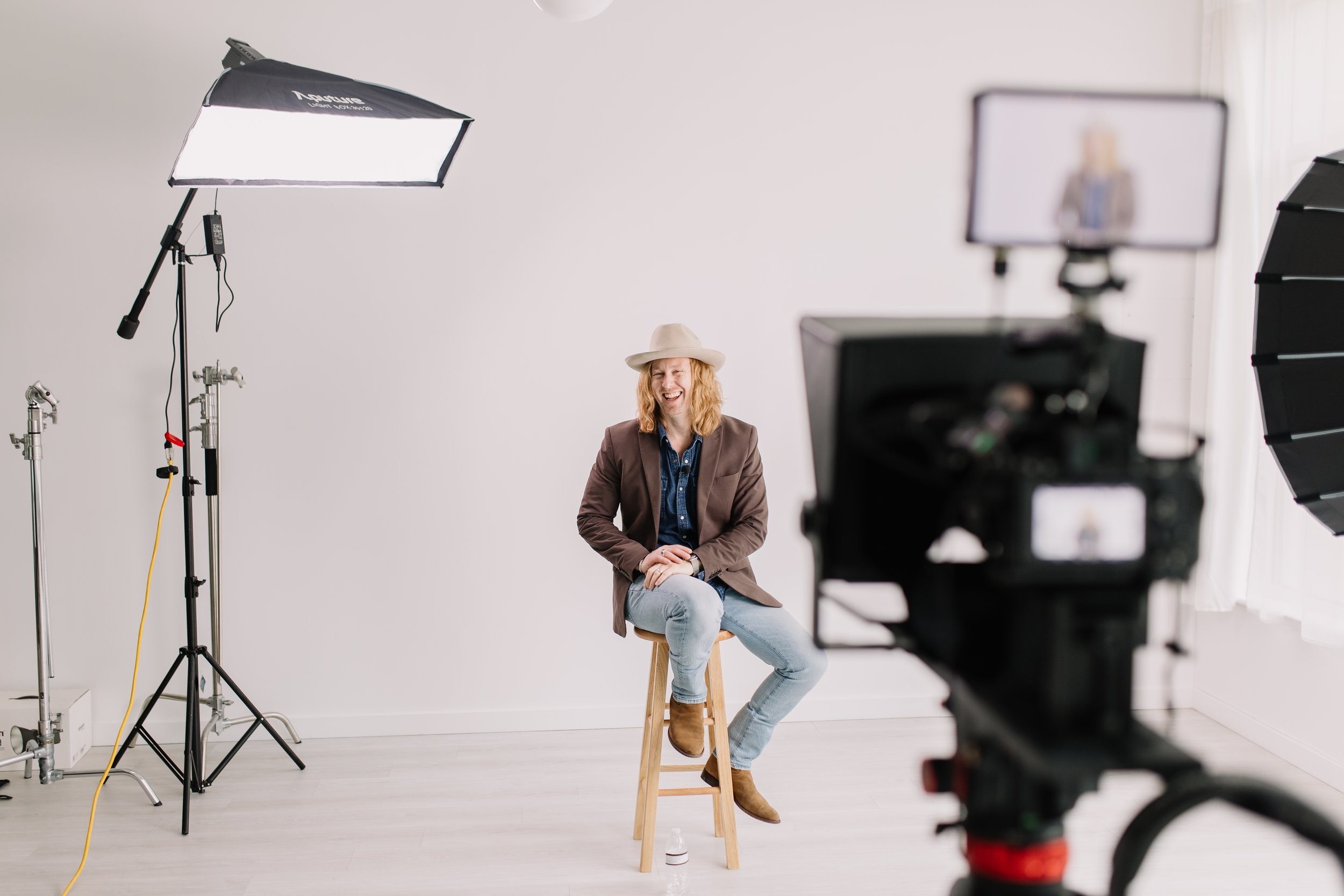 Jamey Ice laughing on a stool in a white studio, a video camera and monitor in the foreground.
