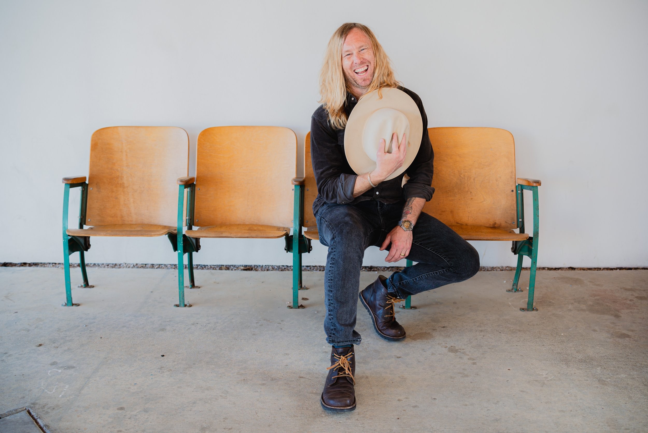 Jamey Ice laughing on a row of old theatre seats, hat in hand