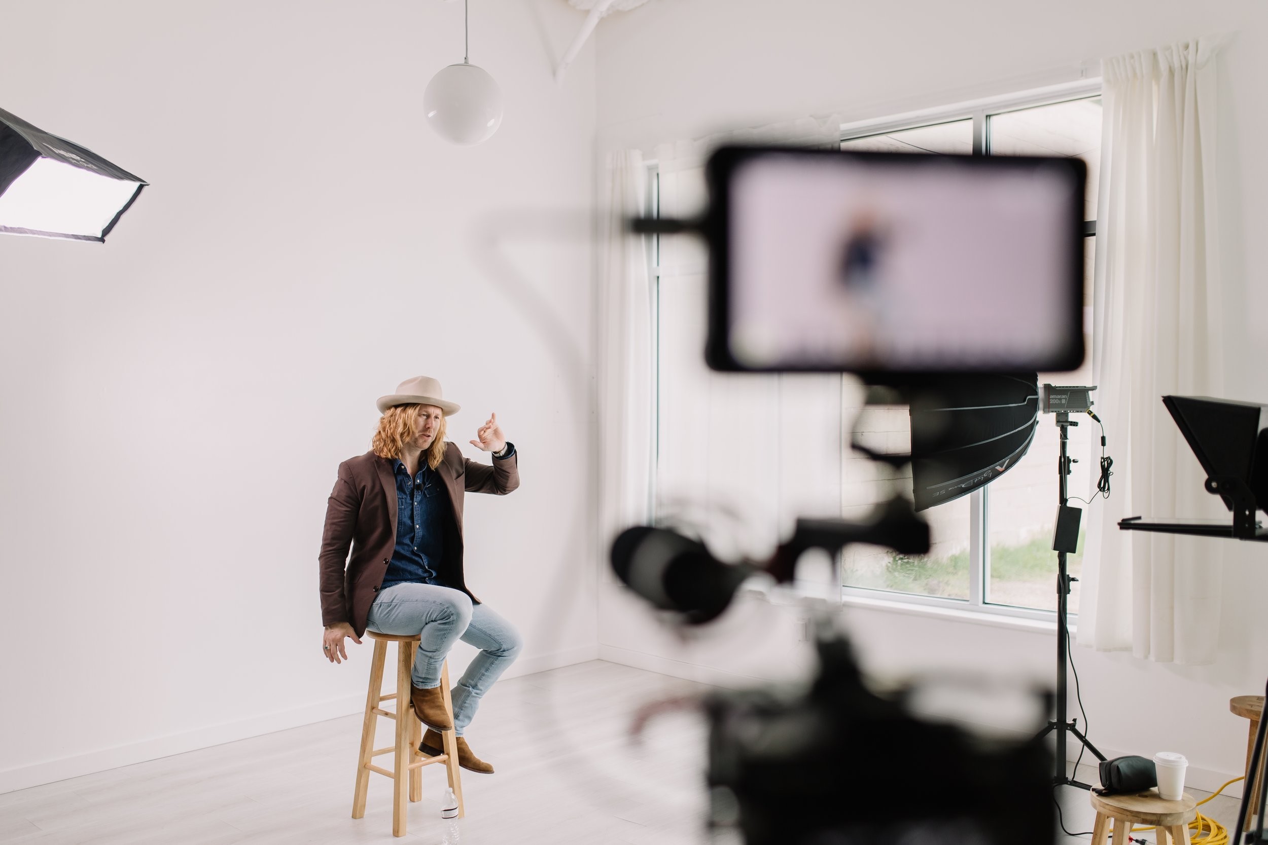Jamey Ice on a stool in a bright studio, a camera on a tripod in the foreground.