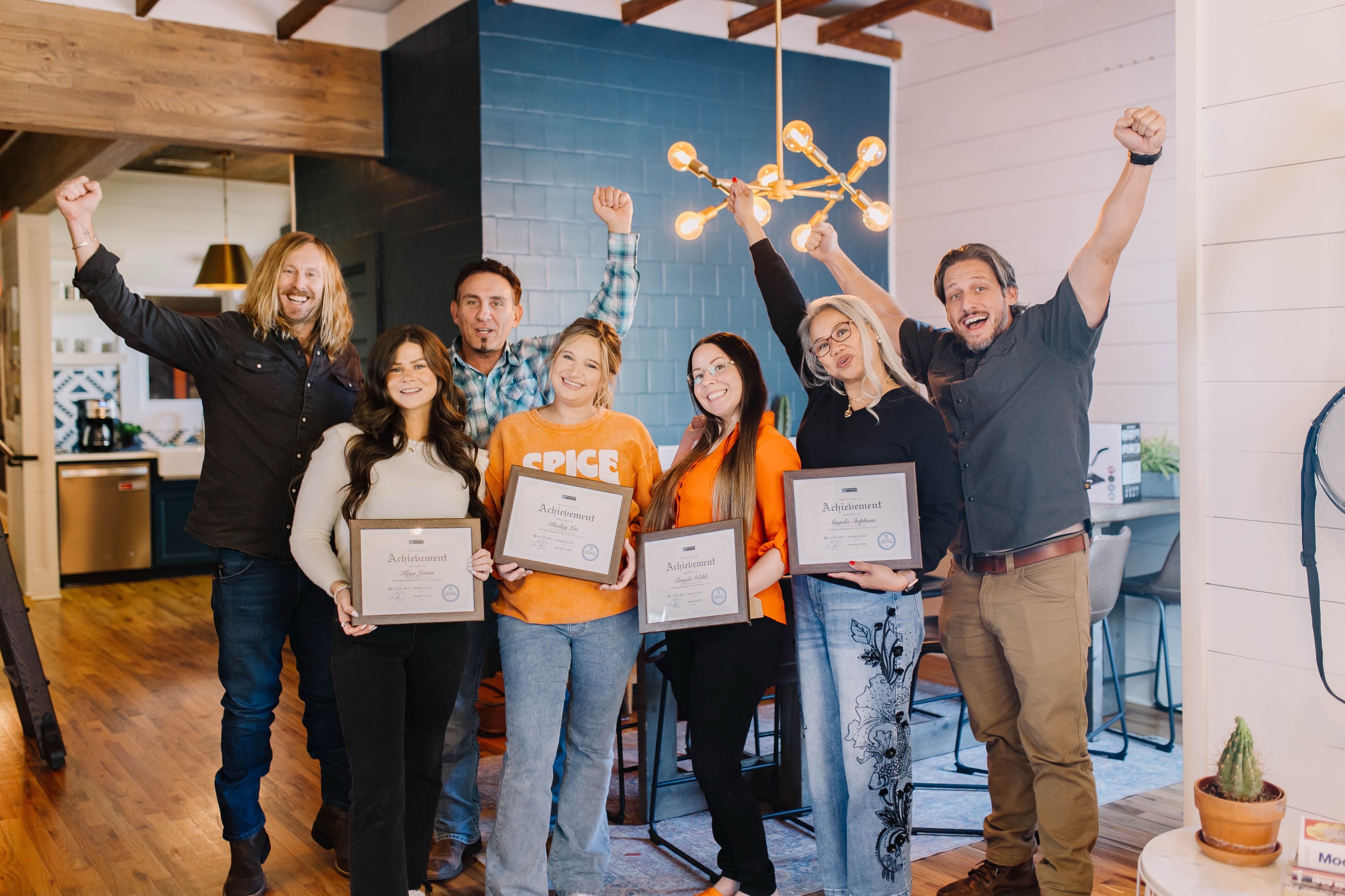 Jamey Ice, one arm up, celebrating with six of the 6th Ave Homes team as the agents hold up their training certificates.