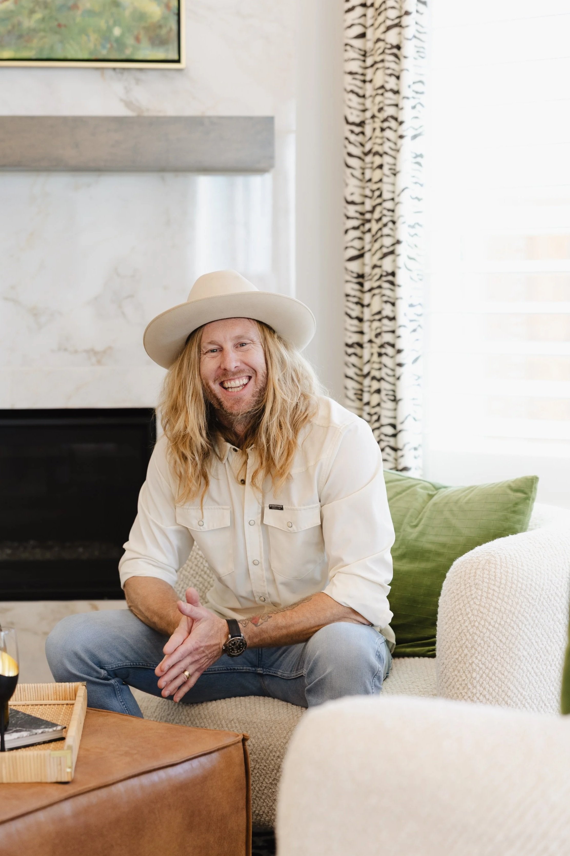 Jamey Ice in a white shirt and hat, seated on a cream couch by a fireplace.