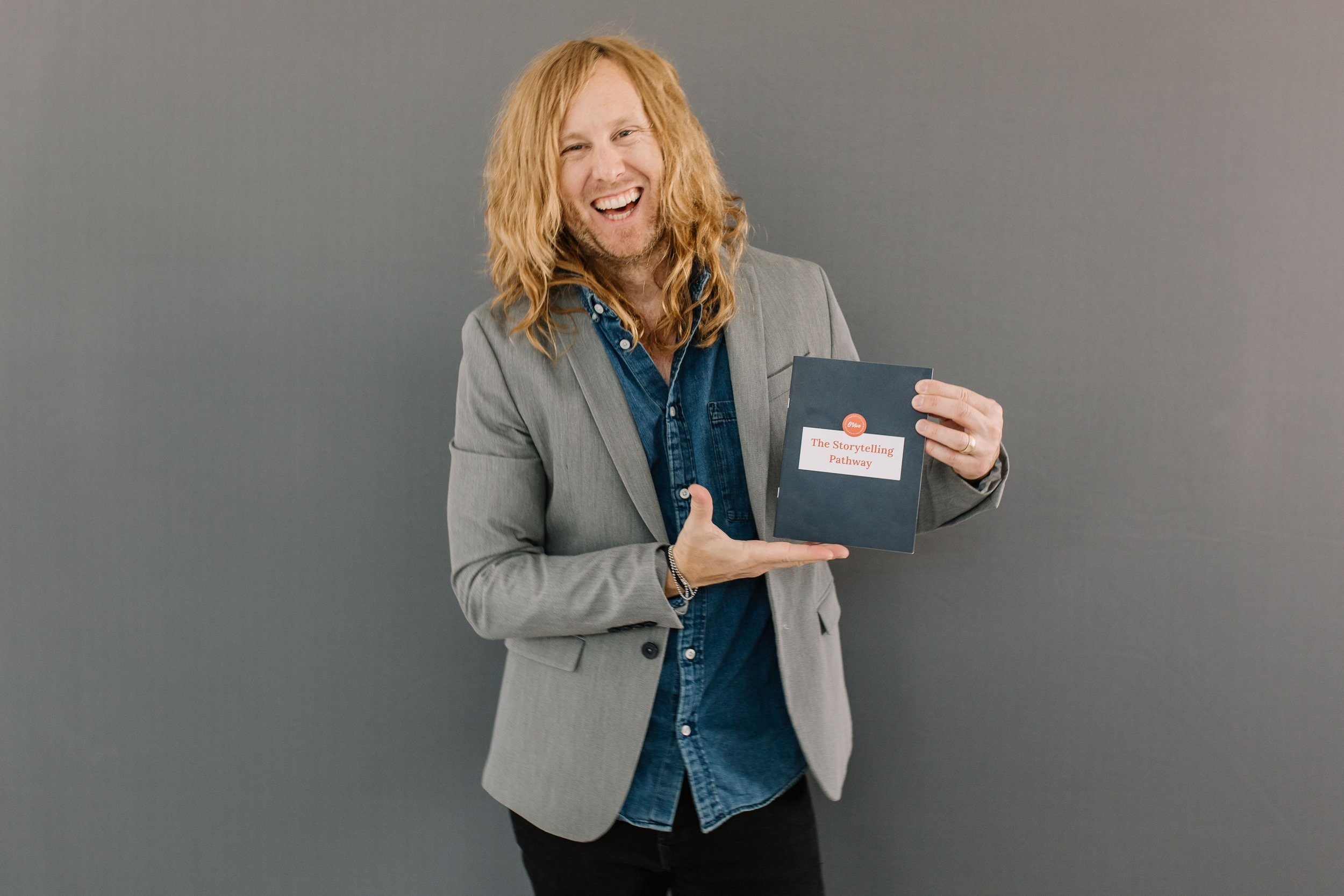 Jamey Ice laughing, holding up a small book against a grey wall.