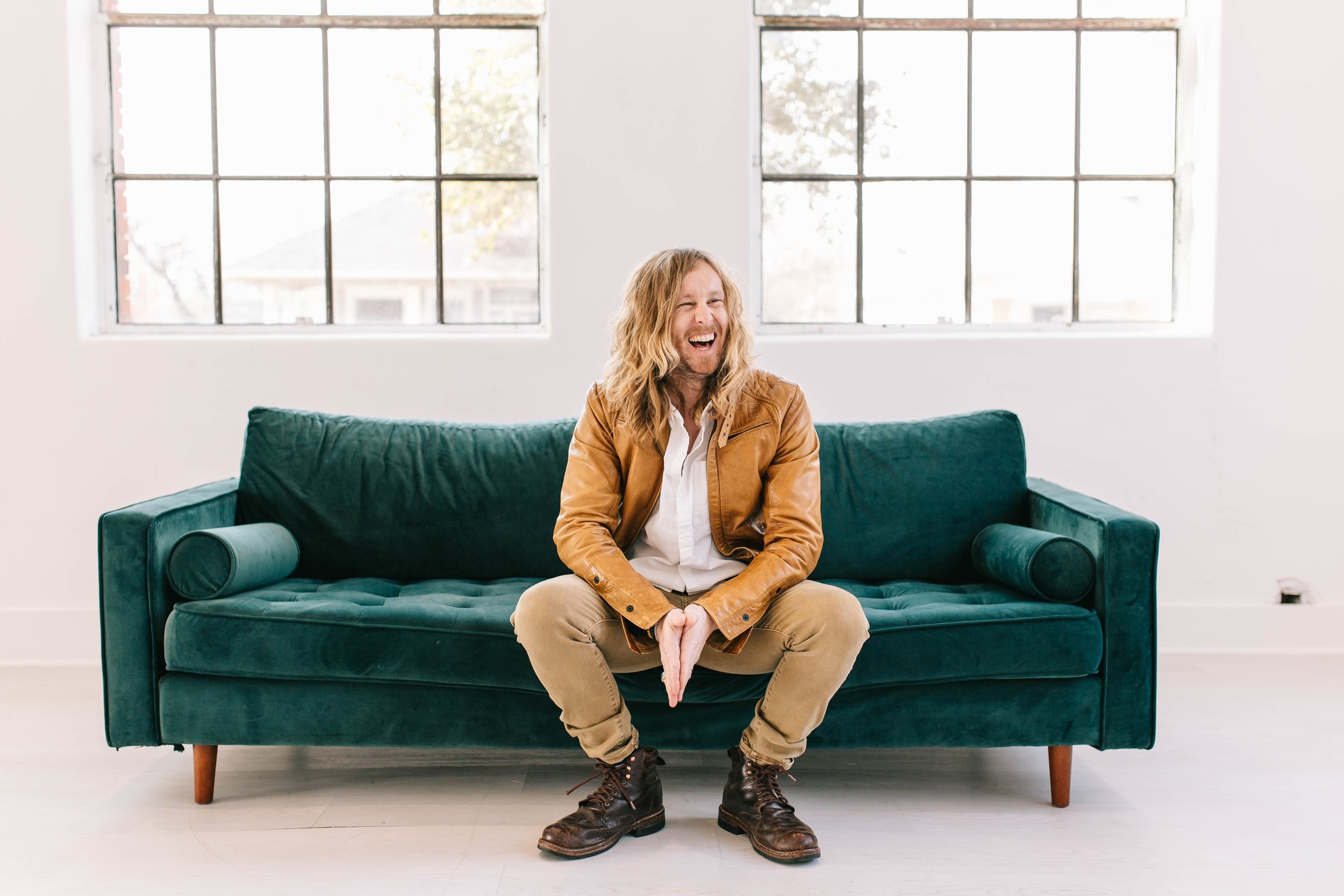 Jamey Ice laughing on a green velvet couch under two tall windows.