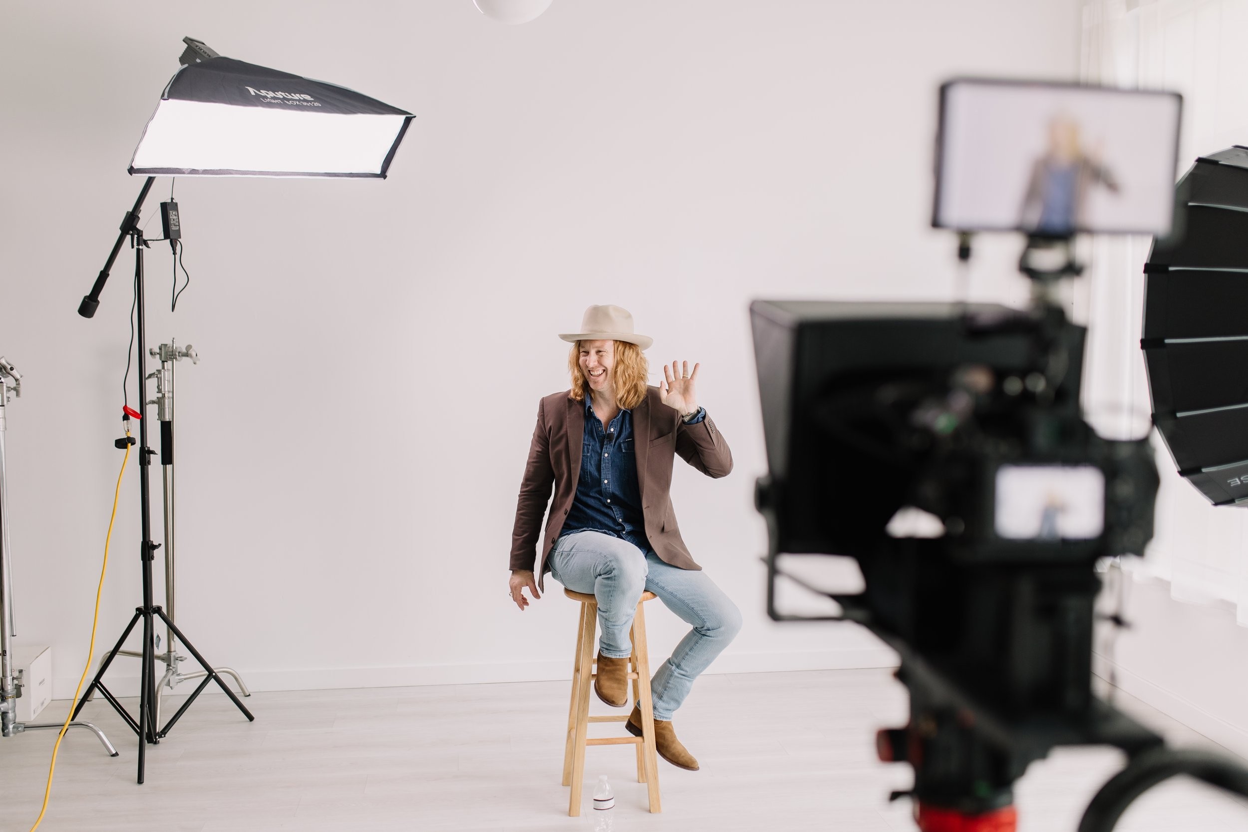 Jamey Ice on a stool in a white studio, waving, a camera and monitor in the foreground.