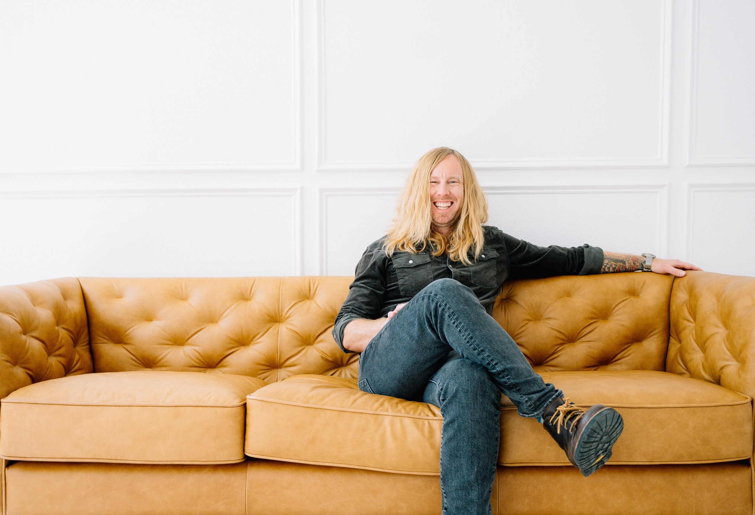 Jamey Ice laughing on a tan leather sofa, one arm along the back