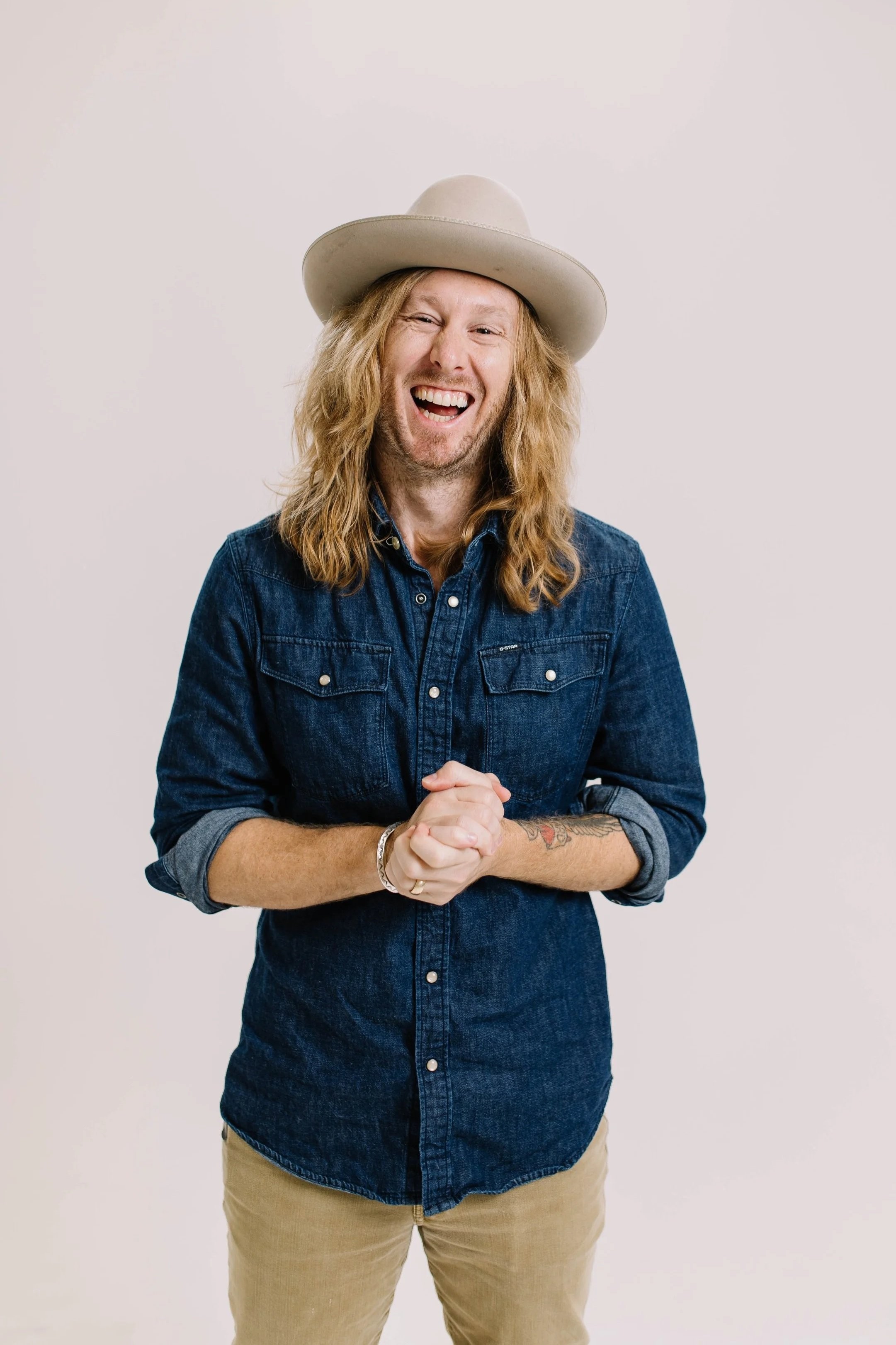 Jamey Ice laughing in a denim shirt and hat, hands clasped, against a pale wall.